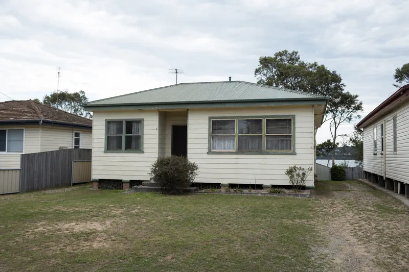 A modest older low-set cottage with a green corrugated roof and a tired front lawn, older houses either side — house demolition, Central Coast. Illustrative example.