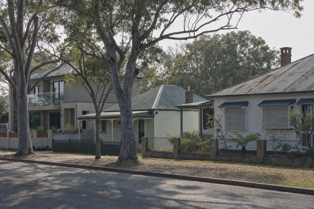 A street where a new two-storey house stands beside older weatherboard cottages with corrugated roofs — house demolition, Central Coast. Illustrative example.