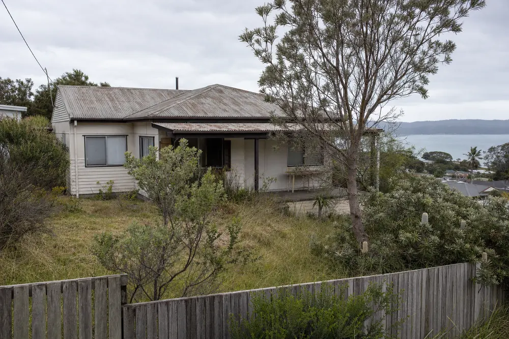 An older single-storey clad house with a corrugated roof on a sloping block, its garden overgrown with long grass and coastal shrubs — house demolition, Central Coast. Illustrative example.