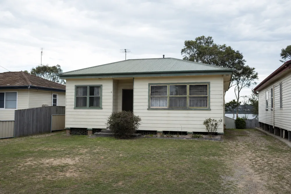 A modest older low-set cottage with a green corrugated roof and a tired front lawn, older houses either side — house demolition, Central Coast. Illustrative example.