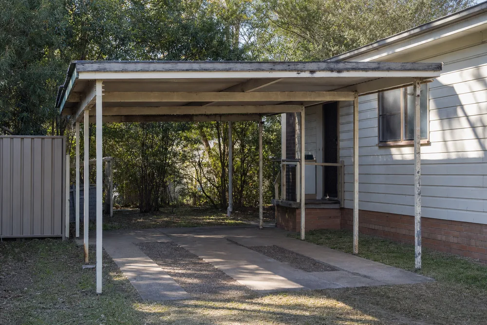 An old flat-roofed carport on painted posts over concrete driveway strips beside a weatherboard house — garage & shed demolition, Central Coast. Illustrative example.